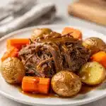 Tender slow cooker pot roast with carrots and potatoes on rustic board.