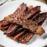 Homemade beef jerky strips with spice rub on a rustic wooden cutting board.